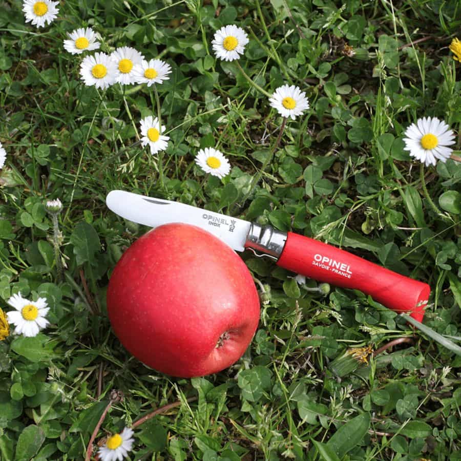 Rød Opinel foldekniv med rustfrit stålblad ved siden af et rødt æble i græs med hvide margueritter